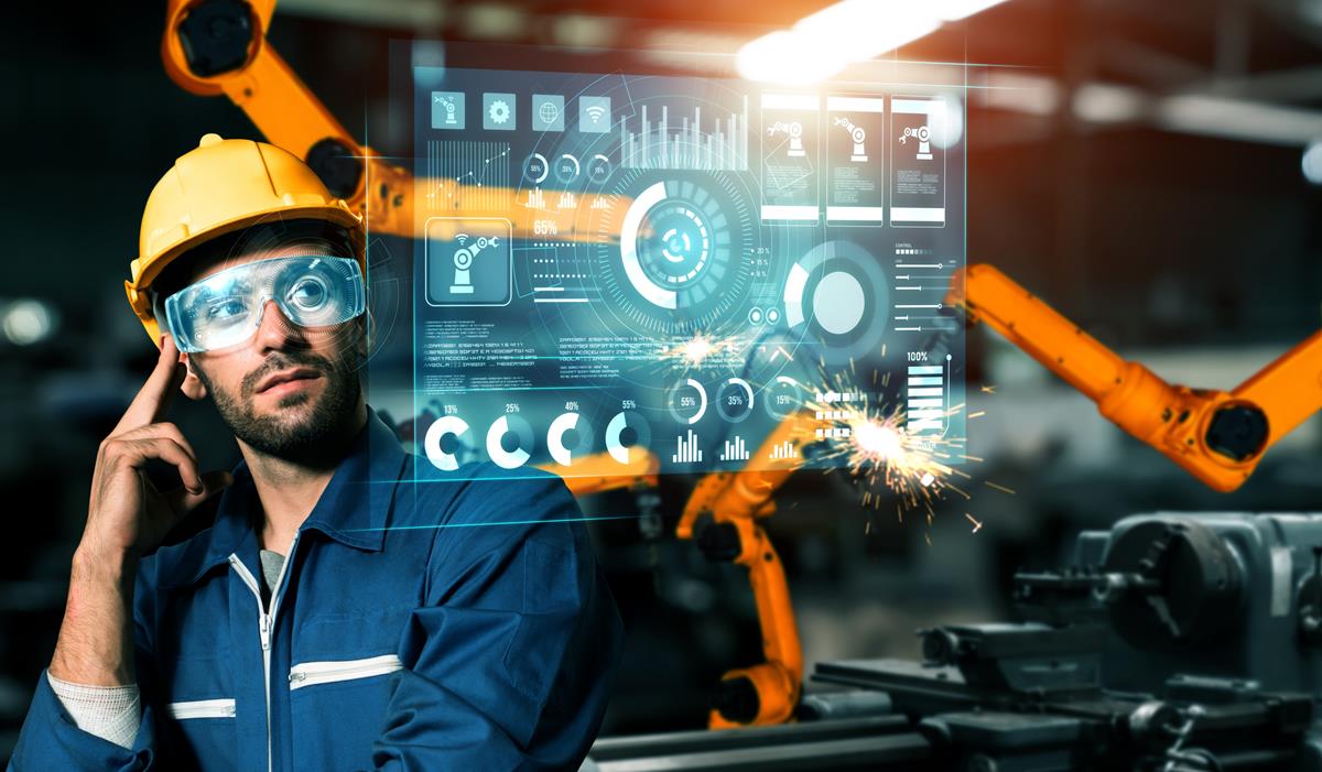 homem vestido de macacão azul, com capacete amarelo, olhando uma tela suspensa, com gráficos.