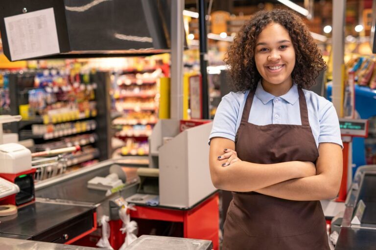 Jovem sorridente caixa em traje de trabalho, cruzando os braços no peito, de trabalho em ambiente de supermercado